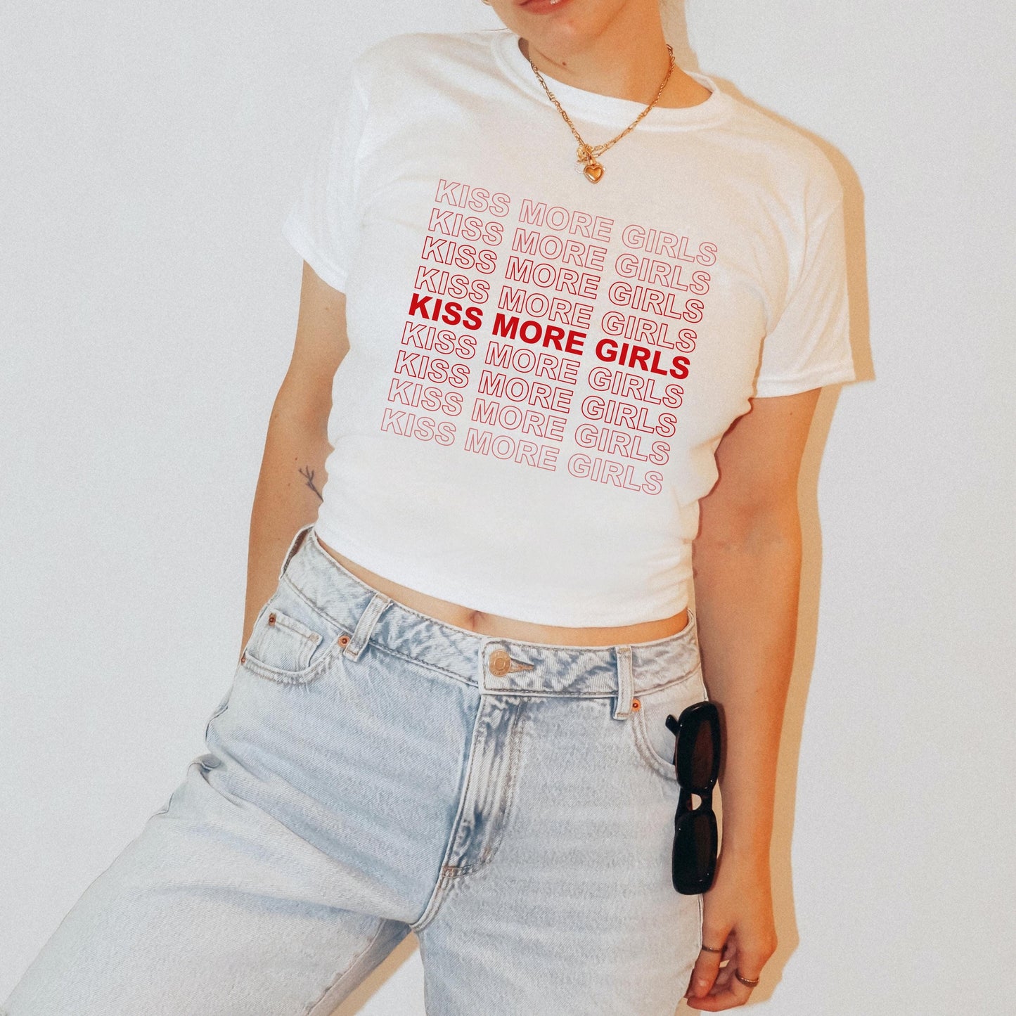 Person wearing a white t-shirt with 'Kiss More Girls' text and light blue jeans on a plain background