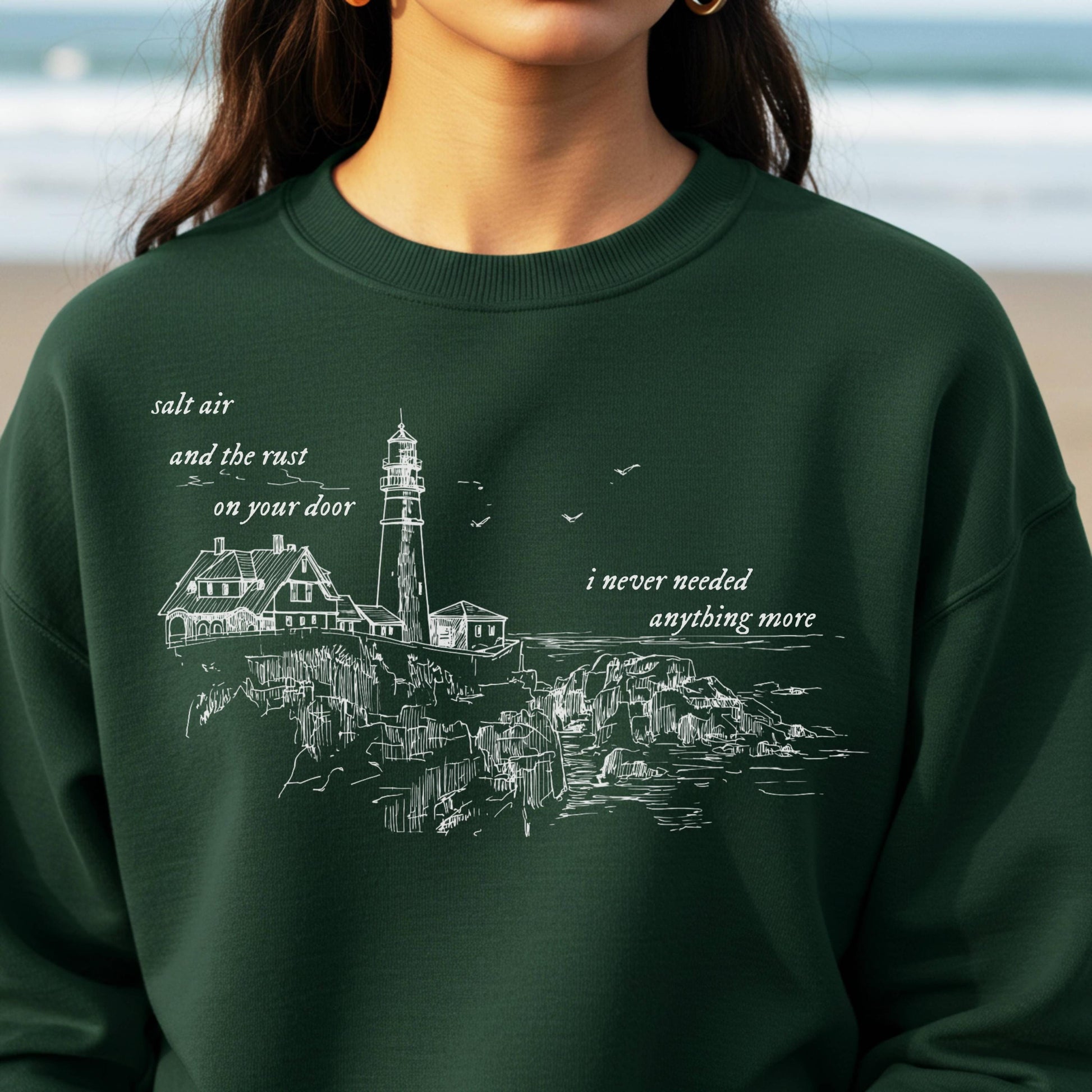 Person wearing a green sweatshirt with a lighthouse design and text on a beach background