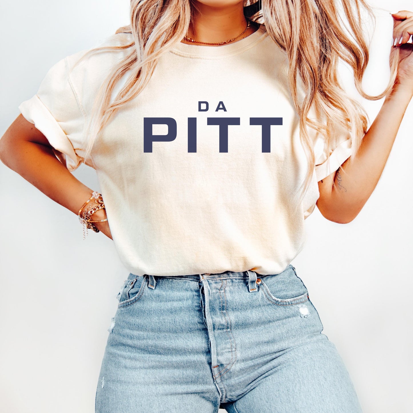 Ivory short-sleeve unisex t-shirt with bold navy lettering reading “DA PITT”. Styled casually with rolled sleeves and light-wash denim jeans.